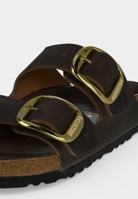 Κοντινό πλάνο από καφέ σανδάλι Birkenstock με δύο δερμάτινους ιμάντες και χρυσοχάλκινες μεταλλικές αγκράφες σε φελλό και μαύρη σόλα.