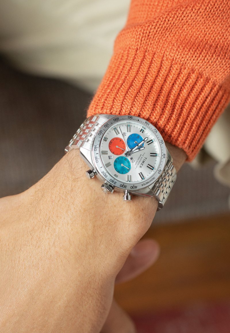 Montre en acier inoxydable argenté avec un cadran blanc, accents rouges, bleus et turquoise, comportant des sous-cadrans et un bracelet en métal tissé.
