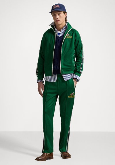 Hanorac verde cu fermoar, cu dungi pe laterale, logo-uri brodate, vestă texturată albastră, cămașă cu dungi, pantaloni verzi asortați și cizme marron.