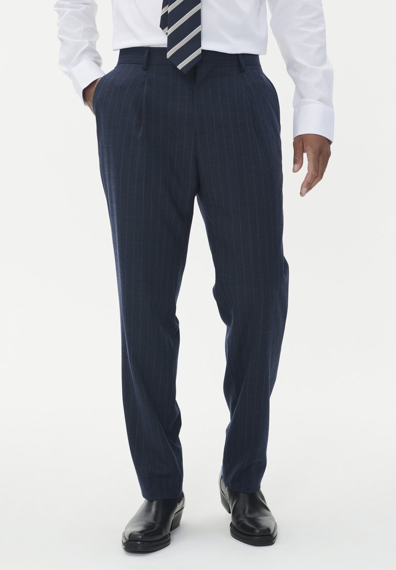 Man som bär marinblå ränderade byxor, vit skjorta, randig slips och svarta dress skor, står med händerna delvis i fickorna.