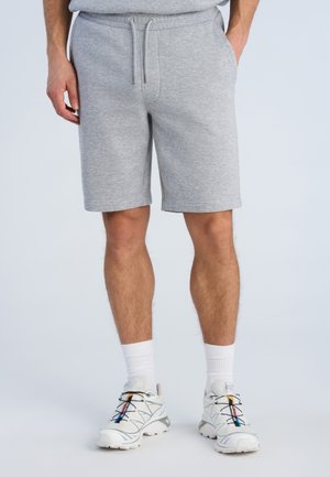 Mężczyzna w jasno szarych szortach z sznurkiem, białych skarpetach sportowych i białych butach sportowych z kolorowymi akcentami, stoi z rękami w kieszeniach.