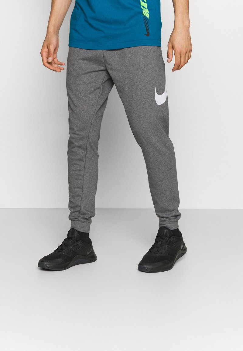 Pantalones deportivos grises de tela suave con un logo blanco de Nike en la pierna izquierda. Combinados con zapatillas deportivas negras y una camiseta azul.