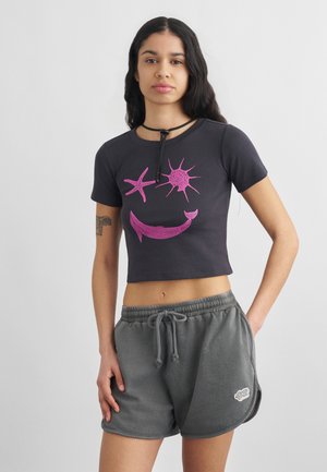 Γυναίκα με μακριά σκούρα μαλλιά φοράει μαύρο κοντό μπλουζάκι με ροζ θαλάσσια πλάσματα και γκρι σορτς με κορδόνι, στέκεται μπροστά σε απλό φόντο.