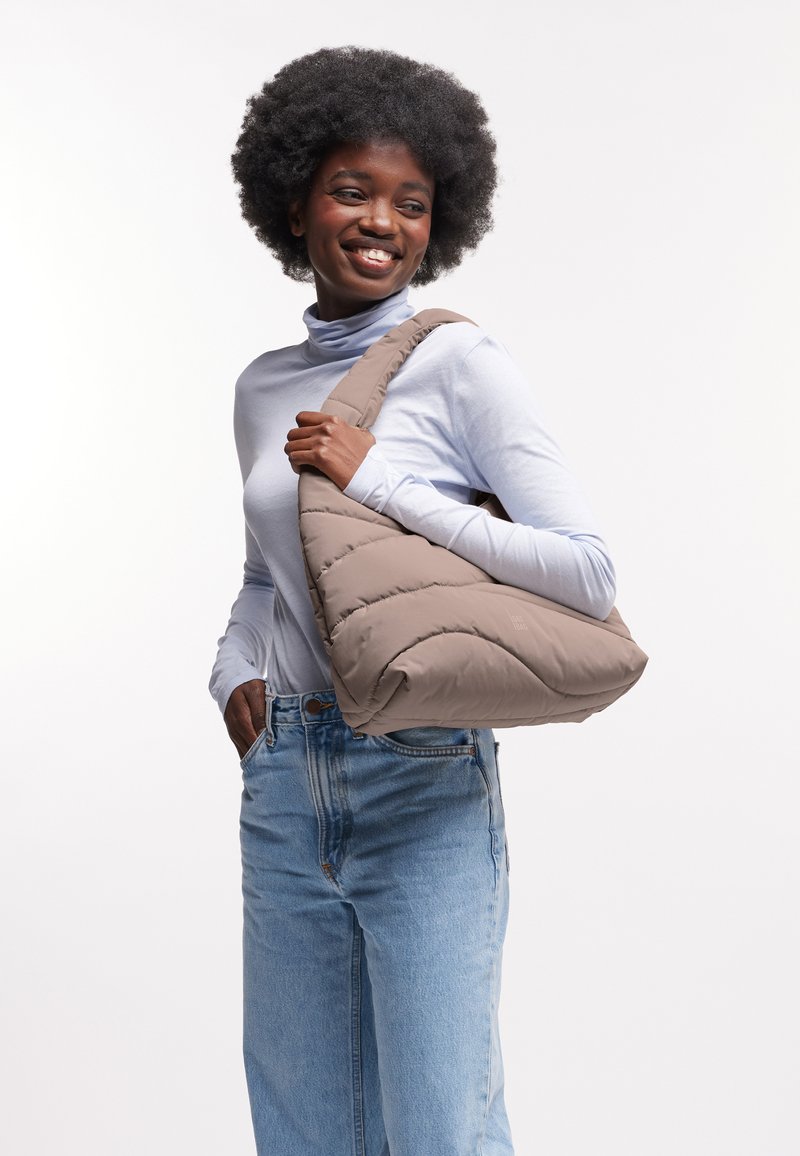 Lachende jonge vrouw met afro-haarstijl, gekleed in een lichtblauwe coltrui en spijkerbroek, met een grote beige gewatteerde schoudertas.