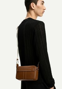 Brown leather crossbody bag with white stitching, rectangular shape, adjustable strap, and a back pocket, worn on a black ribbed sweater.