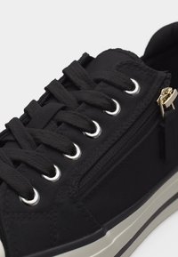 Μαύρο αθλητικό παπούτσι με μαύρα κορδόνια, ασημένιες τρύπες, πλαϊνό φερμουάρ με χρυσό έλασμα και λευκή σόλα με μαύρη ρίγα.