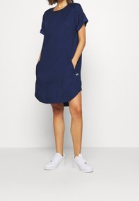 Navyblauwe katoenen jurk met korte mouwen, ronde hals en zijzakken. Heeft een licht gebogen zoom en een klein logo-opdruk.