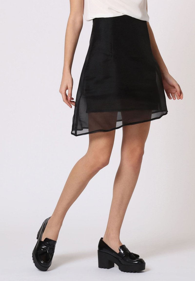 Falda negra asimétrica con un revestimiento transparente, textura suave. Combinada con zapatos de plataforma negros y brillantes, que resaltan el dobladillo.