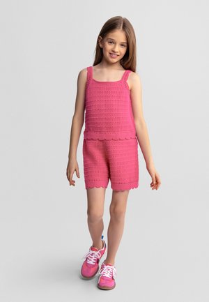 Jong meisje met lang haar, gekleed in een roze gehaakte mouwloze top en bijpassende short, lopend in roze sneakers.