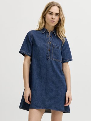 Γυναίκα που φοράει κοντομάνικο μπλε τζιν φόρεμα με μπροστινή τσέπη και κουμπωτό ντεκολτέ, στέκεται μπροστά σε απλό ανοιχτόχρωμο φόντο.