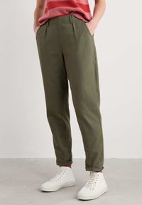 Olivgröna, raka byxor med veck, sidofickor och uppvikta benslut, kombinerade med vita kanvas sneakers och strumpor med röda accenter.