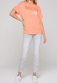 Camiseta corta de manga en color coral naranja con texto blanco y detalles brillantes, combinada con jeans rectos de color gris claro y zapatillas blancas.