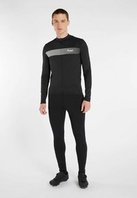 Zwarte sporttop met lange mouwen en een rits aan de voorkant, voorzien van witte horizontale strepen over de borst; gecombineerd met zwarte aansluitende leggings en zwarte schoenen.