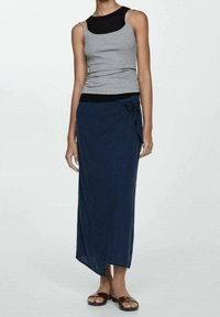 Graues Tanktop und dunkelblauer Wickelrock mit einer Schleifendetail an der Taille. Der Rock ist knöchellang mit einem asymmetrischen Saum und wird mit braunen Sandalen kombiniert.