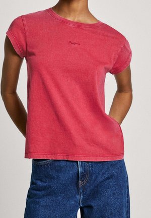 Personne portant un t-shirt rouge délavé à manches courtes légèrement rentré dans un jean bleu taille haute, debout avec une main dans la poche.