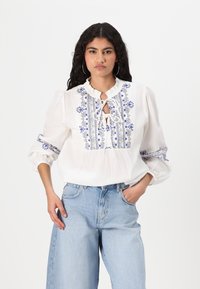 Blusa bianca ampia realizzata in tessuto leggero, con ricami floreali blu, colletto arricciato e dettaglio con lacci al collo.