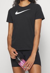 Camiseta negra de algodón con mangas cortas y un gran logo blanco de Nike en el pecho, combinada con pantalones cortos negros; sosteniendo una botella de agua rosa.