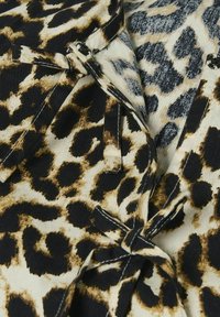 Leoparda raksta audums bēšā un melnā krāsā ar tekstūras dziļumu, ar saitēm un kontrastējošiem šuvju detalizējumiem ap kakla līniju.