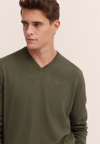 Olive green, long-sleeve V-neck sweater made of soft fabric, featuring a subtle logo detail on the chest and a relaxed fit.