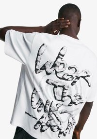 Bílý bavlněný tričko s výrazným černým grafickým textovým designem na zádech. Krátké rukávy, oversized střih a jemná textura.