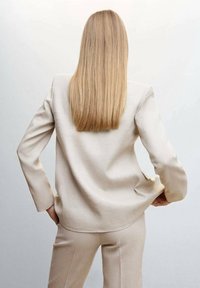 Béžový přizpůsobený blazer s dlouhými rukávy, hladkým povrchem a rozparkem na zadní straně. Doplněný o odpovídající béžové kalhoty.