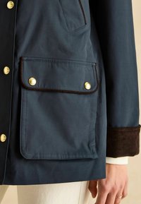 Jachetă impermeabilă de marină cu nasturi din bronz, manșetă din catifea și o buzunar mare cu tiv cusut și detaliu de branding.