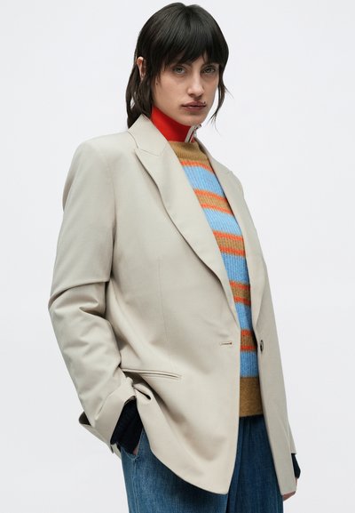 Μπεζ σακάκι με μοτίβο, με γιακά με αιχμή, κλείσιμο με ένα κουμπί και πλαϊνές τσέπες. Φορεμένο πάνω από πολύχρωμο ριγέ πουλόβερ και τζιν παντελόνι.