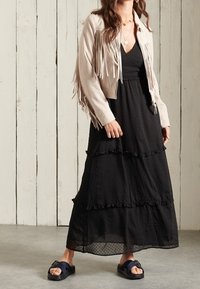 Helle beige Lederjacke mit Fransen-Details über einem schwarzen, gestuften Maxikleid mit Punkten. Schwarze Rutschen-Sandalen runden das Outfit ab.