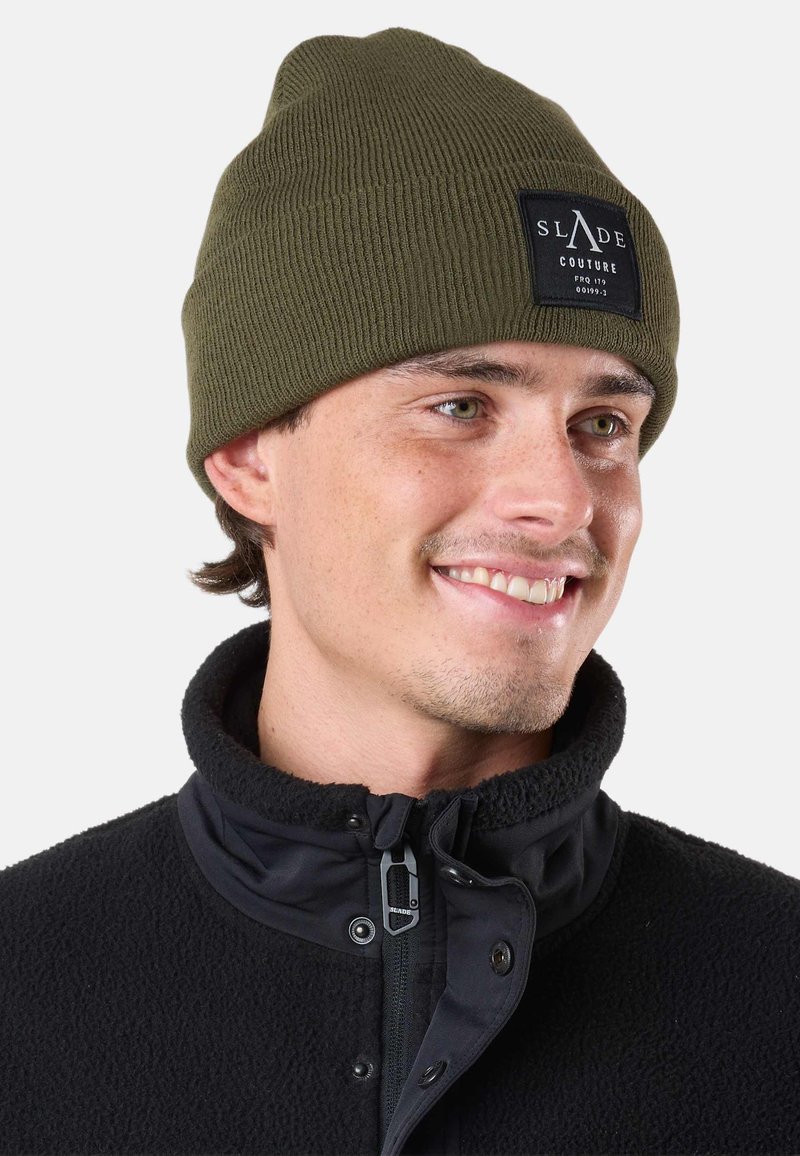 Jeune homme souriant, portant un bonnet olive Slade Couture et une veste polaire noire avec une fermeture éclair et des boutons-pression.