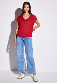 Czerwony sweter typu V-neck z krótkim rękawem, zestawiony z jasnoniebieskimi spodniami o szerokich nogawkach i białymi trampkami. Cienie nadają głębi.