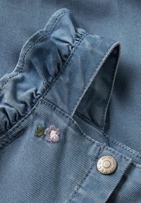 Denimstoff med lys blå nyanse, prydet med en rynket halslinning, blomsterbroderi i lilla og grønt, og en sølvknapp med tekst.