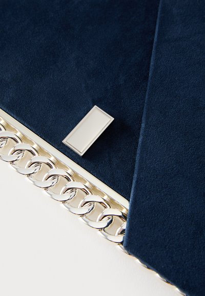 Tamsiai mėlynas zomšos rankinukas su sidabrine grandinės detalė ir stačiakampiu metaliniu ženkleliu. Turi lygią tekstūrą ir tvarkingą dizainą.