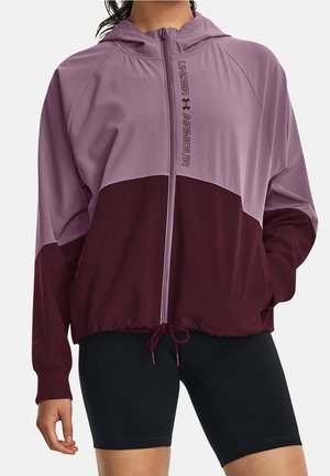 Frau trägt eine zweifarbige lila Under Armour Zip-Up-Jacke mit Kapuze und schwarze Radsport-Shorts, steht mit einer Hand in der Tasche.