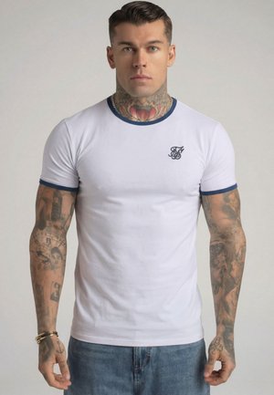 Mand med tatoveringer på nakke og arme iført en tætsiddende hvid T-shirt med blå kant og blå jeans, stående mod en ensfarvet baggrund.