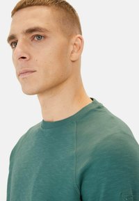 Green crew-neck sweatshirt with textured fabric, raglan sleeves, and a subtle logo patch on the left sleeve. Smooth finish, casual design.