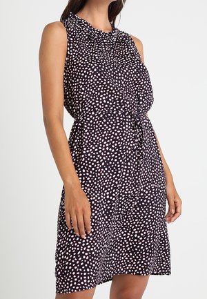 Černé šaty bez rukávů s růžovými puntíky, nařaseným výstřihem a páskem v pase, které má na sobě stojící žena na jednobarevném pozadí.