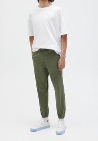 Camiseta blanca de gran tamaño, pantalones jogger verde oliva con cordón ajustable y zapatillas blancas con suelas translúcidas azules.