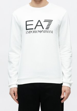 Osoba ubrana w białą koszulę z długim rękawem z czarnym logo "EA7 Emporio Armani" i czarne spodnie z małym białym logo po lewej stronie.