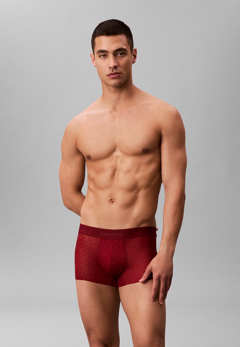 Czerwone bokserki Calvin Klein z siateczkową strukturą, z logo na pasku i geometrycznym wzorem, noszone przez męski model.