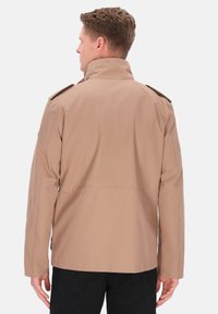 Beige jakke med høj krave, strukturerede skuldre og to knaplukninger på epauletterne; glat stof og ren syning langs sømmene.