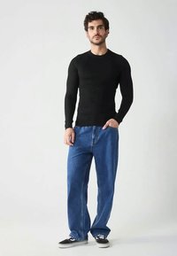 Giovane uomo in piedi, con una maglietta nera a maniche lunghe aderente, jeans blu larghi e sneakers bianco e nere, di fronte a uno sfondo chiaro e semplice.