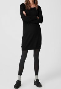 Donna in piedi con le braccia incrociate, indossa un vestito nero a maniche lunghe, collant neri, calze grigie e sneakers nere su uno sfondo neutro.