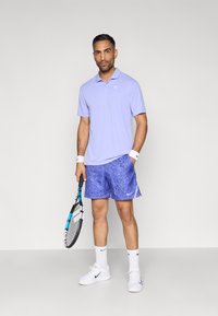 Ανοιχτό μωβ κοντομάνικο πόλο με γιακά και λογότυπο της Nike, συνδυασμένο με μπλε σορτς με σχέδια και λευκά παπούτσια του τένις, που φοριέται από έναν άνδρα με ρακέτα.