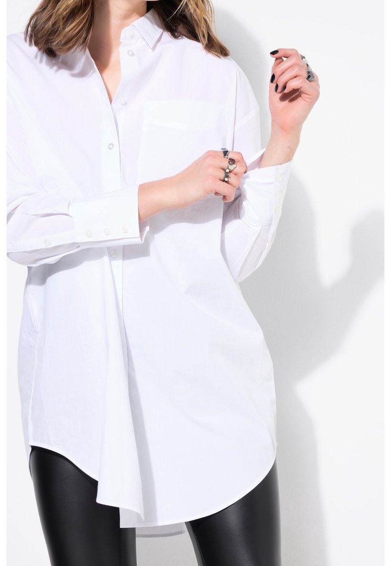 Personne ajustant le poignet d'une longue chemise blanche à boutons, portant des bagues noires et un pantalon en cuir noir sur fond blanc.