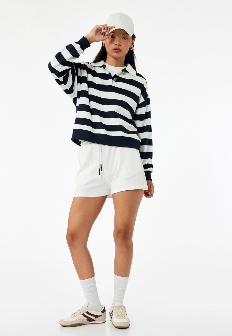 Gestreepte marineblauwe en witte langemouwige polo, witte shorts, witte pet, witte sokken, casual sneakers met mahoniekleurige accenten.