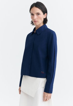 Donna con capelli corti e scuri che indossa una camicia blu navy con bottoni e una gonna bianca asimmetrica su sfondo bianco.