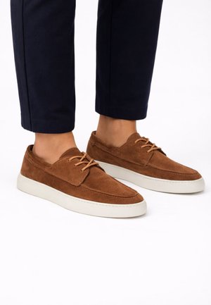 Braune Wildleder-Schnürschuhe für die Freizeit, getragen zu schwarzen, knöchellangen Hosen, vor weißem Hintergrund.