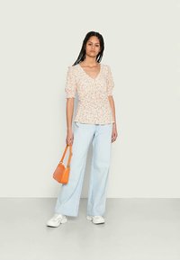 Blusa floral clara com mangas puff, decote em V e bainha peplum, combinada com jeans de perna larga azul claro e uma mala laranja. Ténis brancos.