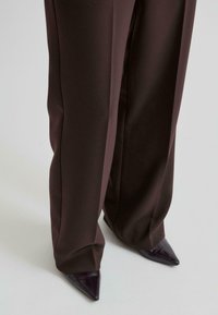 Pantalon large marron foncé porté sur des chaussures en cuir marron foncé à bout pointu sur un sol gris clair.