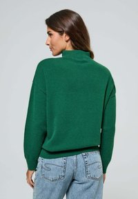 Pull en tricot vert avec un col montant, coupe décontractée et épaules tombantes. Associé à un jean taille haute bleu clair.